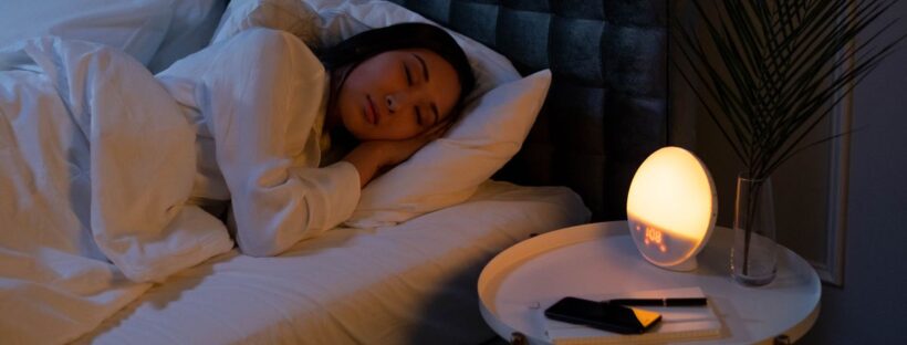 A person sleeping peacefully in a dark bedroom lit by a warm, low bedside lamp